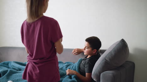 rear view of young girl handing over tea to her sick brother lying on the couch, who reaches out to collect it and drink it, showing sibling affection and care