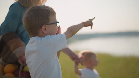 a close-up of a young boy wearing glasses and a white shirt, pointing at the sky with excitement, while his mother holds his younger sibling in the background near a peaceful lake