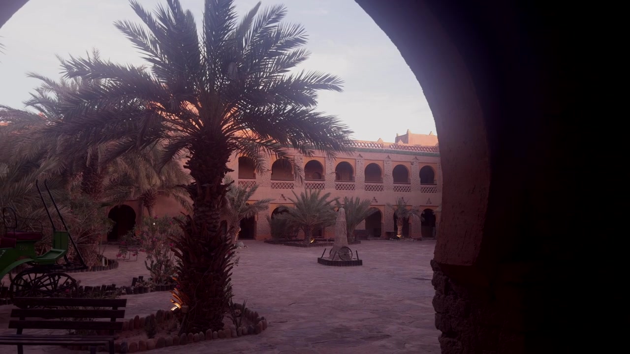 Grande cortile di un hotel kasbah con fontana centrale, palme e balconi ad arco a due piani al tramonto, Marocco.