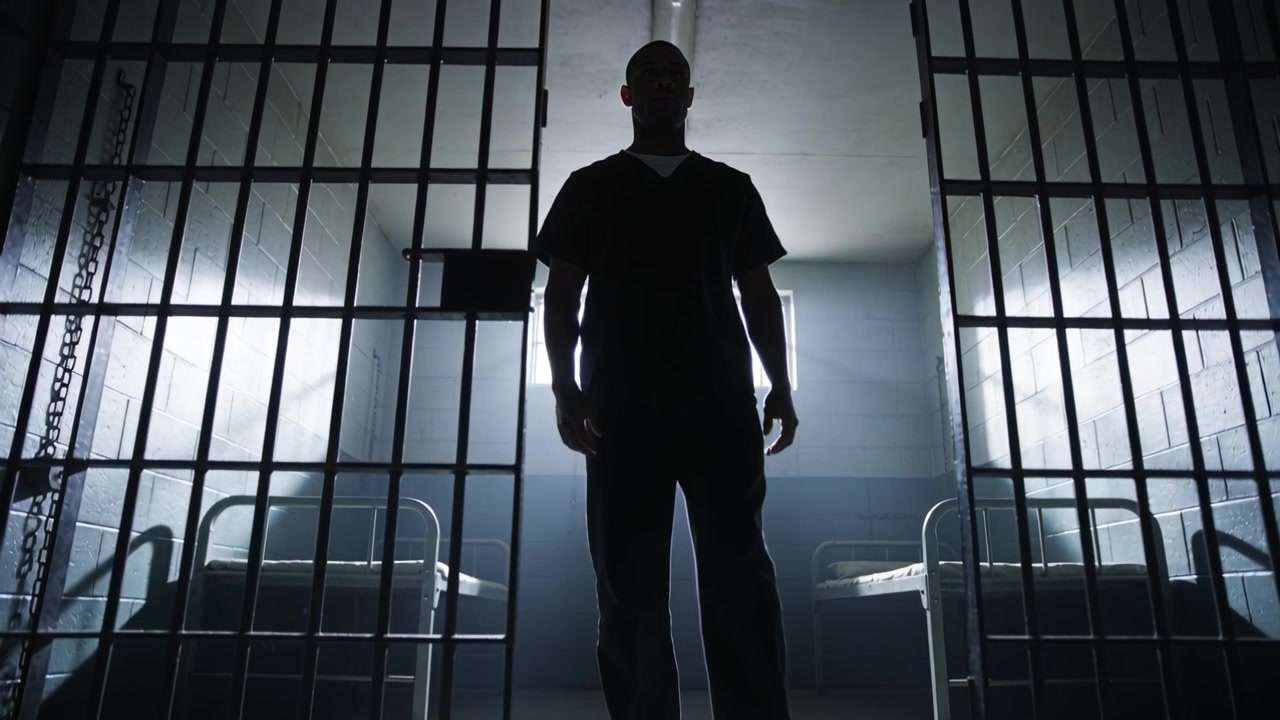 Un detenuto maschio adulto che avanza dalla porta sbarrata di una cella, indossando l'uniforme e con letti a castello.