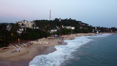 Luftdrohne-über-Dem-Touristischen-Reiseziel-Am-Sandstrand,-Das-Mit-Menschen,-Hunden-Und-Sonnenschirmen-Bei-Sonnenuntergang-In-Sri-Lanka-Beschäftigt-Ist