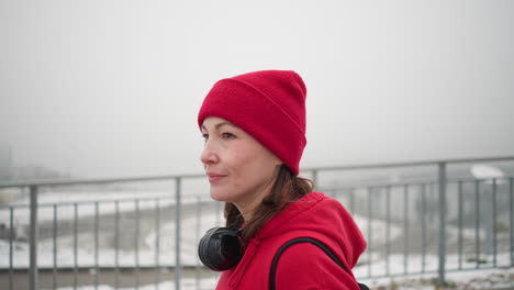 close-up of woman walking with a subtle smile wearing red beanie, headphones around neck, near iron railing, background shows blurred view of bridge and urban settlement in foggy atmosphere