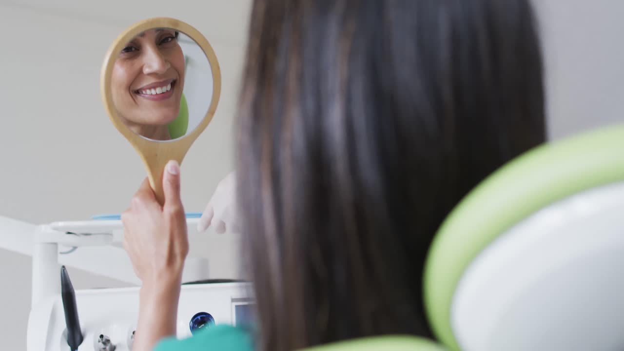 Biracial female patient looking at teeth and smiling in mirror at modern dental clinic