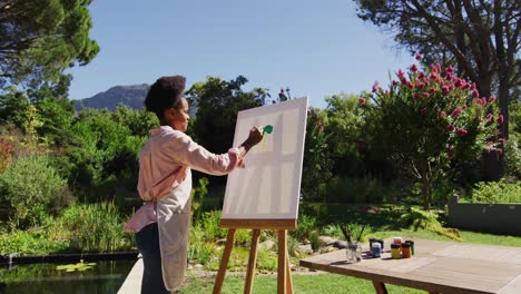 smiling african american woman painting picture on canvas in sunny garden