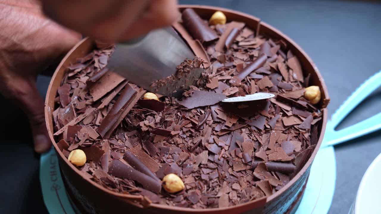 Hands slicing a deluxe chocolate cake topped with nuts, shavings, and a raspberry—perfect for elegant food visuals