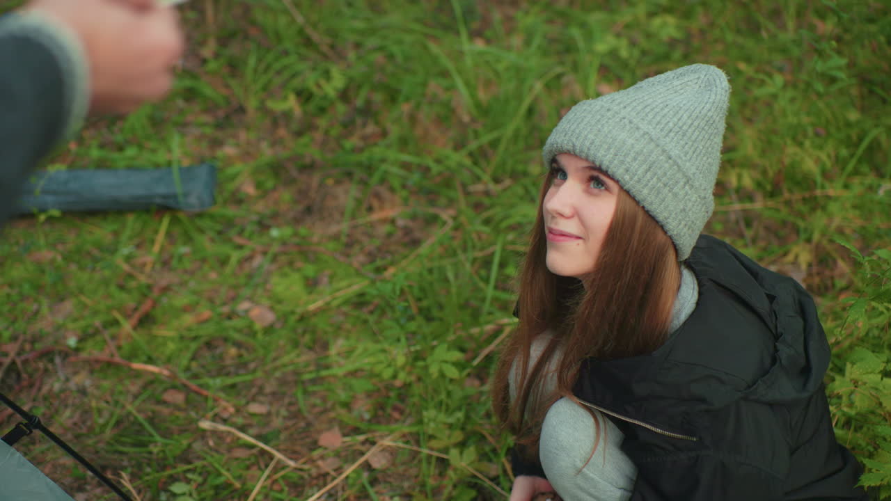 Young woman in warm outdoor clothing squats on grassy forest ground, gazing affectionately at man in front of her while reaching out to collect tent peg to assist in setting up camping shelter