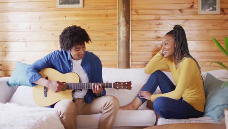 happy african american couple playing guitar, sitting on sofa in log cabin, slow motion