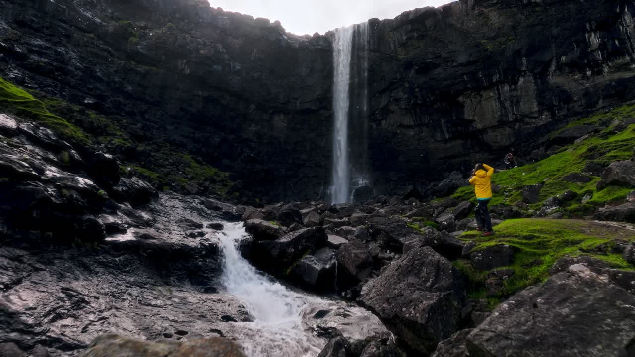 un turista con una chaqueta amarilla fotografía una majestuosa cascada de las islas feroe al aire libre
