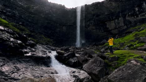 a tourist in a yellow jacket photographs a majestic faroe islands waterfall outdoors