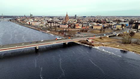 Antena-Del-Puente-De-La-Carretera-Que-Abarca-Un-Ancho-Río-Azul-Daugava-En-Una-Ciudad-Densamente-Poblada-De-Riga-Con-Altos-Rascacielos-En-El-Fondo,-Toma-Aérea-En-órbita