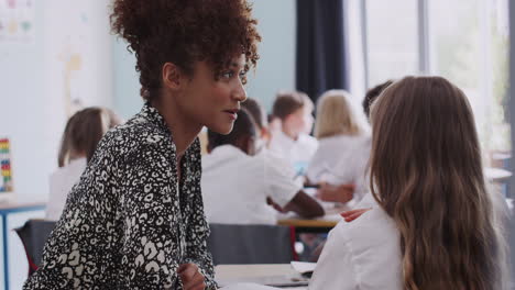 Woman-Elementary-School-Teacher-Giving-Female-Pupil-Wearing-Uniform-One-To-One-Support-In-Classroom