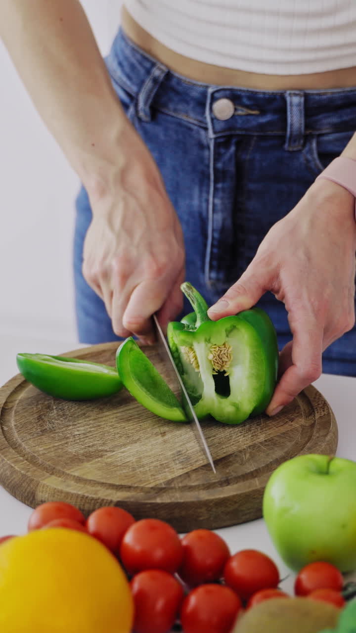 Young female is cutting green pepper on a wooden board. Close-up woman hands preparing healthy food on the kitchen on fresh ingredients background. Vertical video