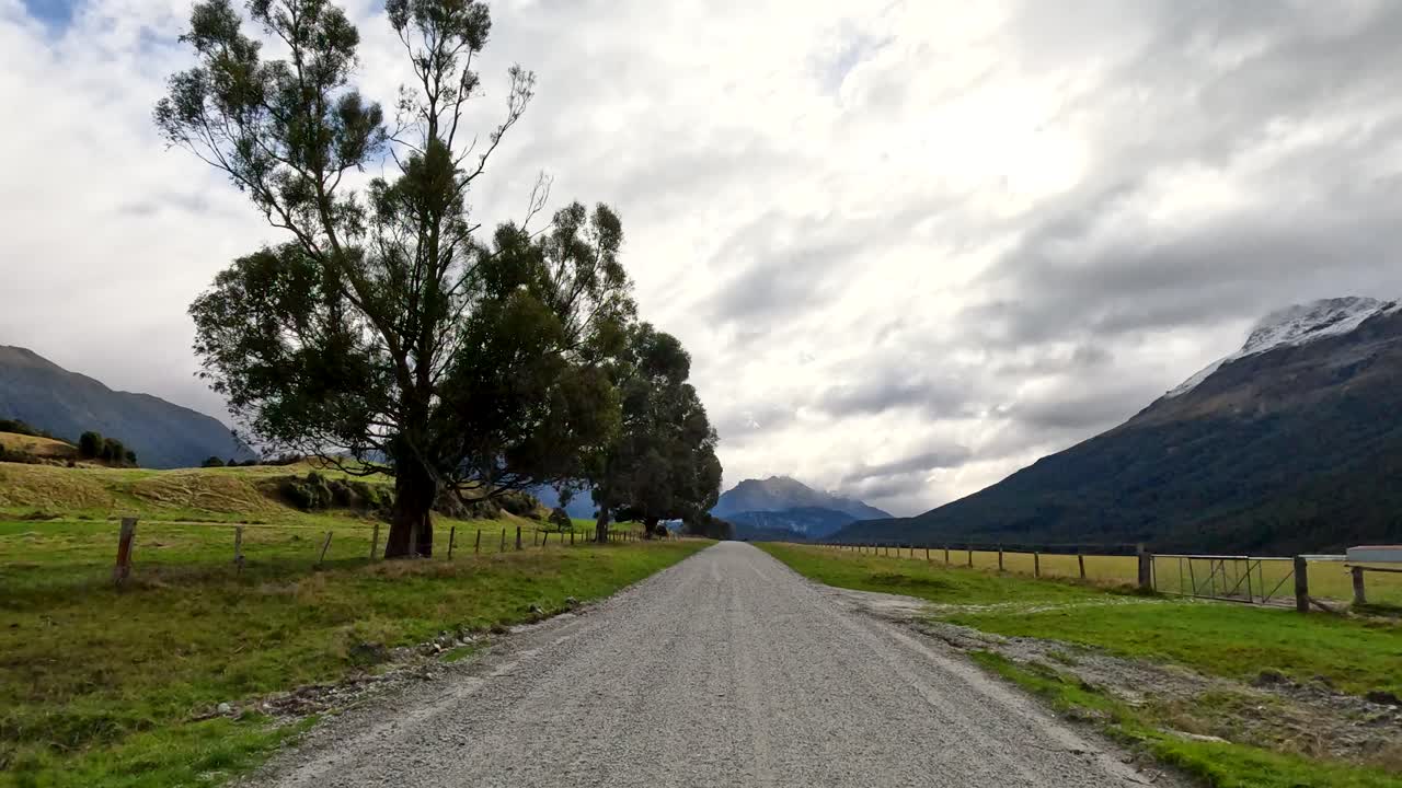 Vehicle moves along rural gravel road, passing trees, with overcast sky and mountain backdrop