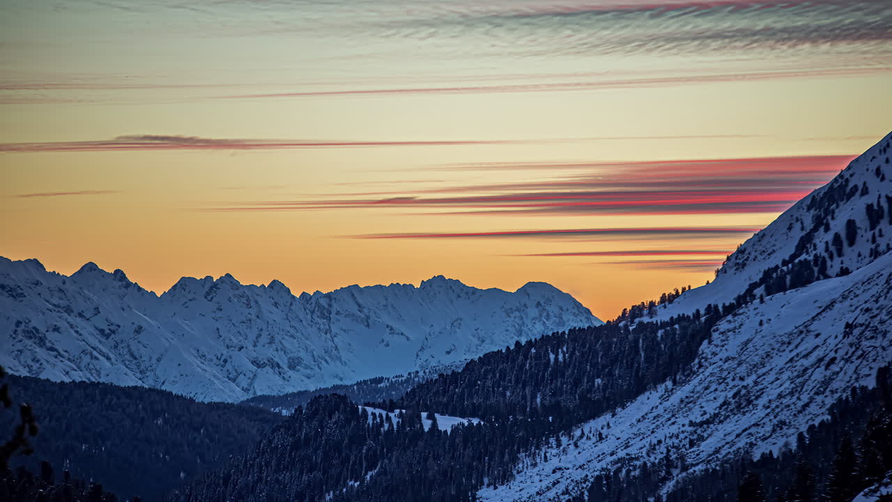 Timelapse: Tramonto arancione, nuvole in volo e cime innevate con foresta di pini durante il tramonto.