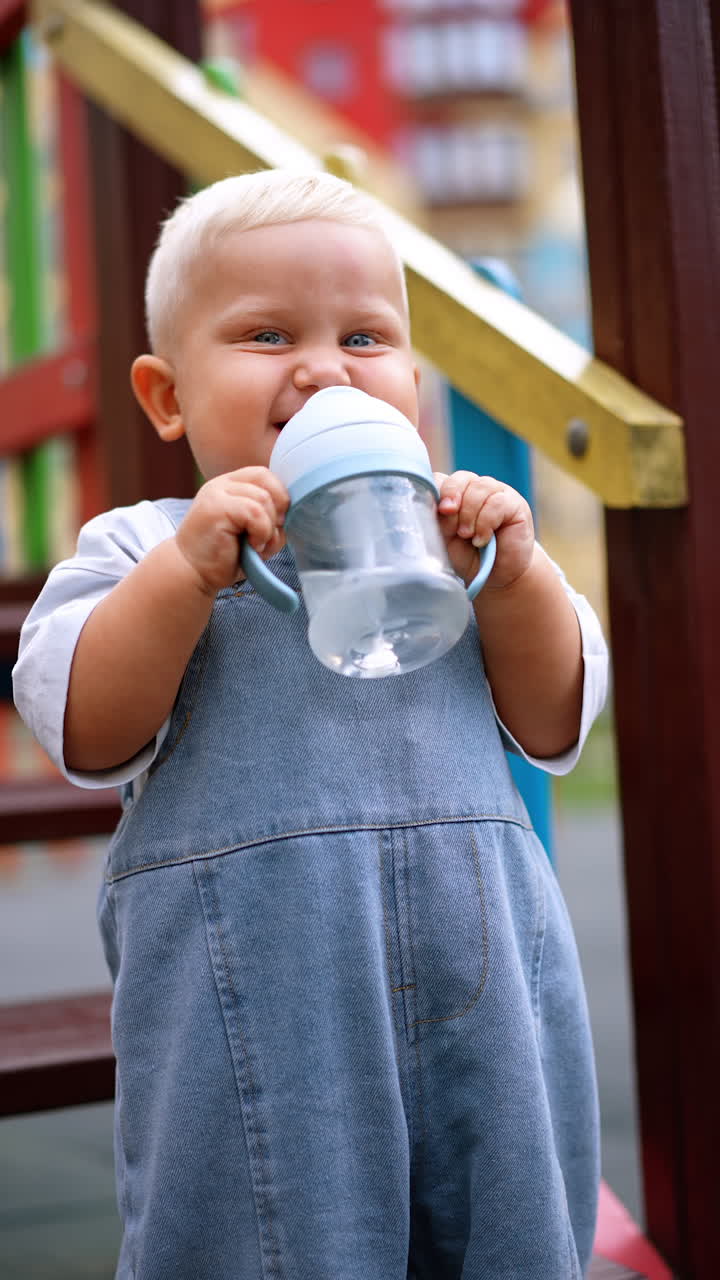 Adorable blond baby in jeans romper drink water from bottle and smiles. Lovely toddler dances and waves his hand. Vertical video.