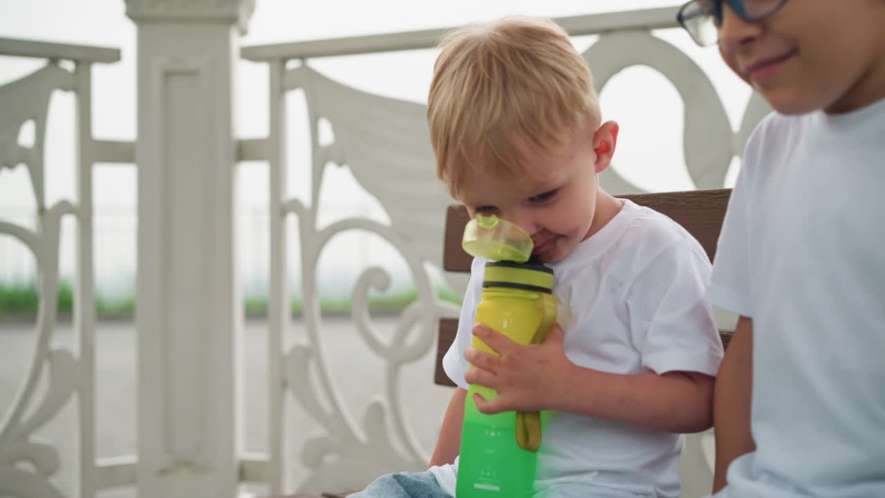 dos hermanos jóvenes están sentados juntos, el niño más joven tiene una botella de agua verde cerca de su boca, preparándose para beber