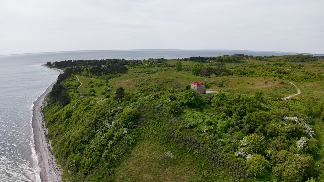 Aerial view of a lush green headland along a steep coastline, featuring a small red-roofed building and winding path above the sea