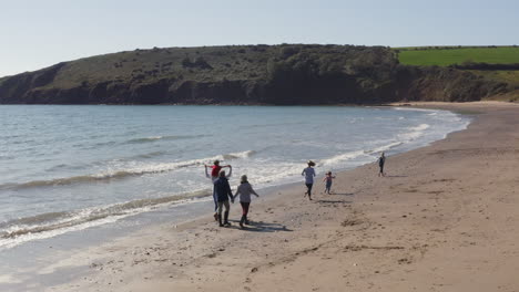 Drohnenaufnahme-Einer-Familie-Mit-Mehreren-Generationen-Im-Urlaub,-Die-Am-Strand-Entlang-Spaziert-Und-Wellen-Bricht