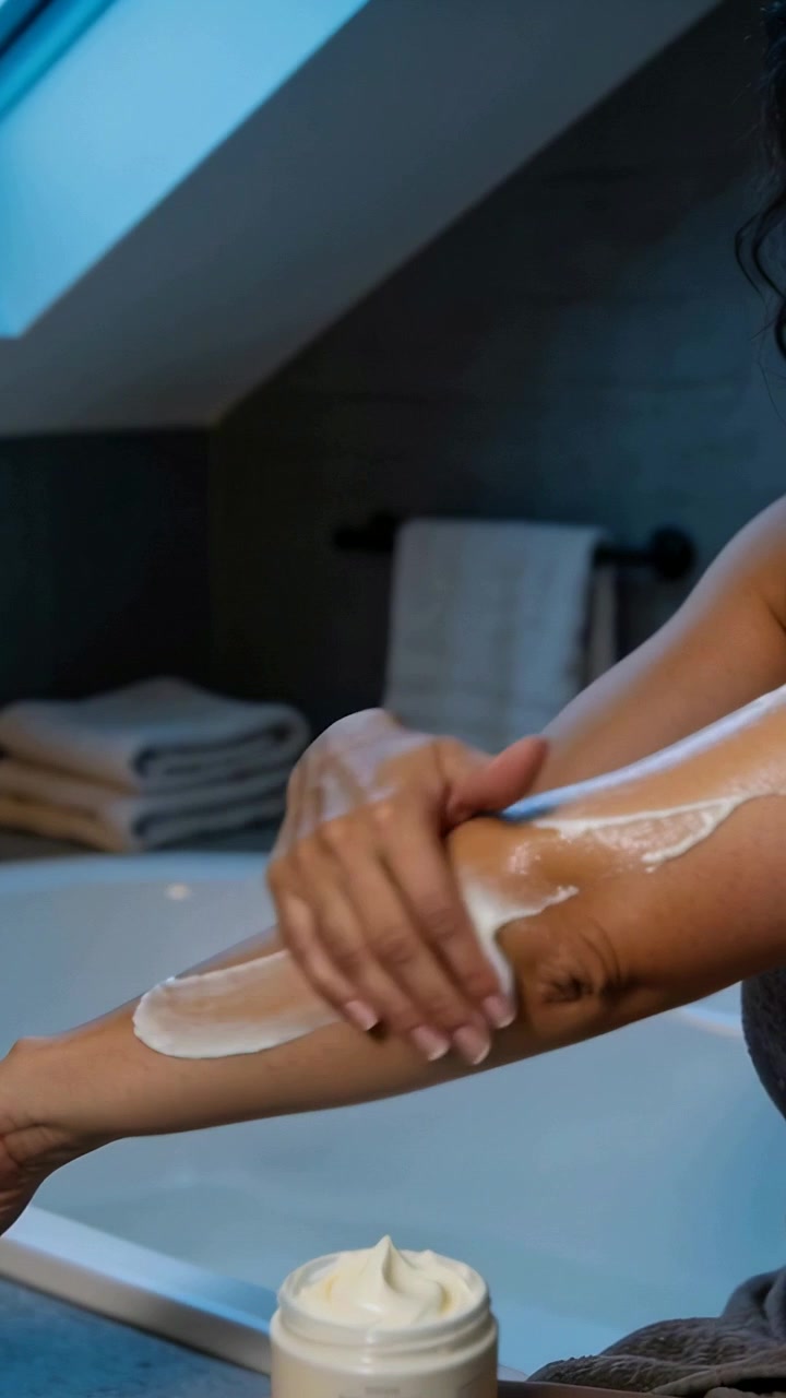 Video verticale: Donna avvolta in un telo da bagno che preleva la crema e si strofina gli avambracci per idratarli vicino alla vasca, con un vasetto