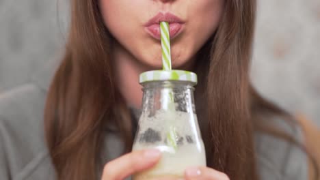 close-up scene of a beautiful girl who is sitting at home on the couch, drinking a homemade milkshake from a bottle with a straw