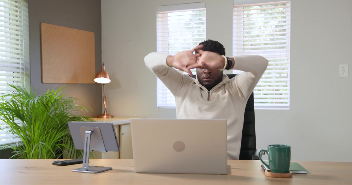 Homme afro-américain faisant une pause dans son bureau à domicile, étirant son cou devant son ordinateur portable pour soulager la tension avant de reprendre le travail.