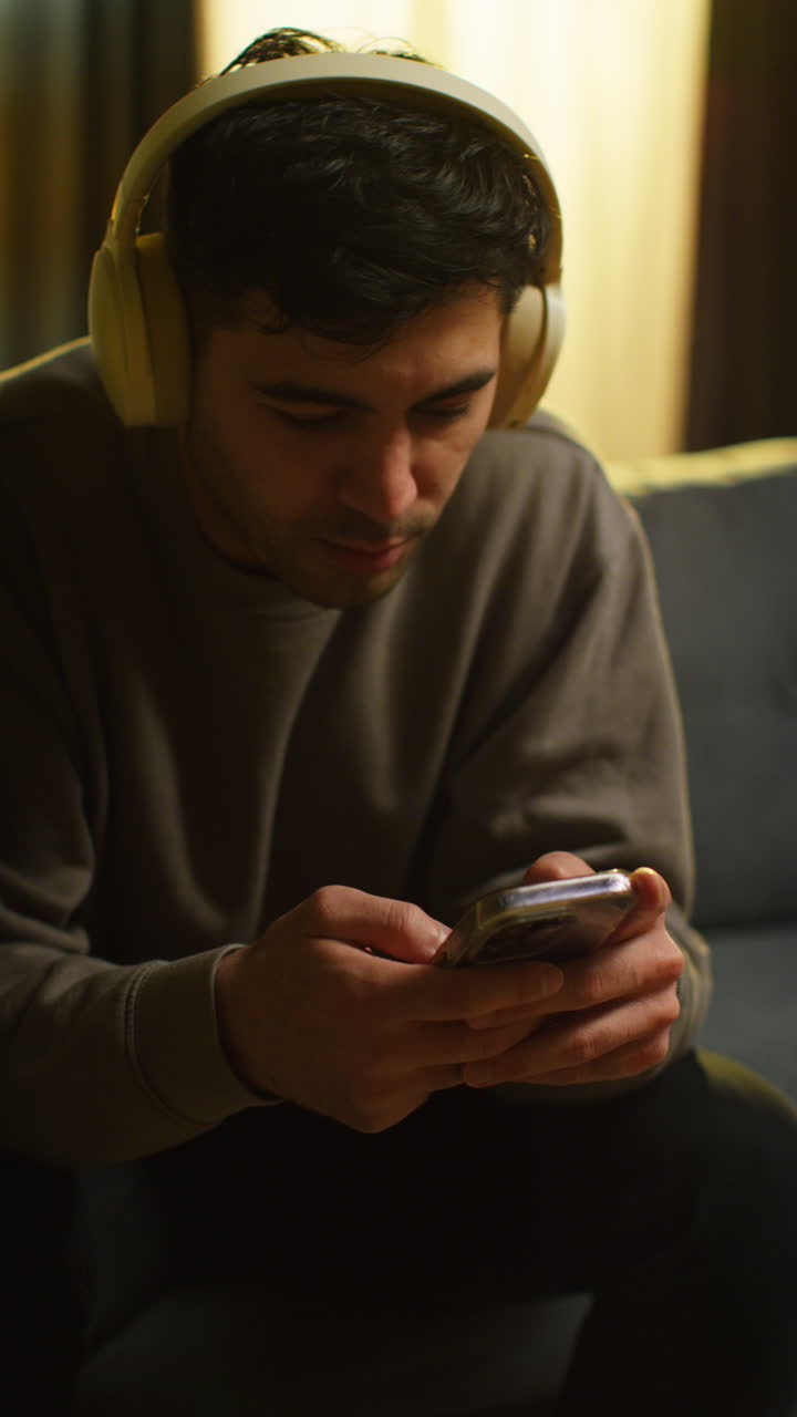 Vertical Video Of Man Spending Evening At Home Sitting On Sofa Wearing Wireless Headphones Streaming Music Or Watching Content From Mobile Phone