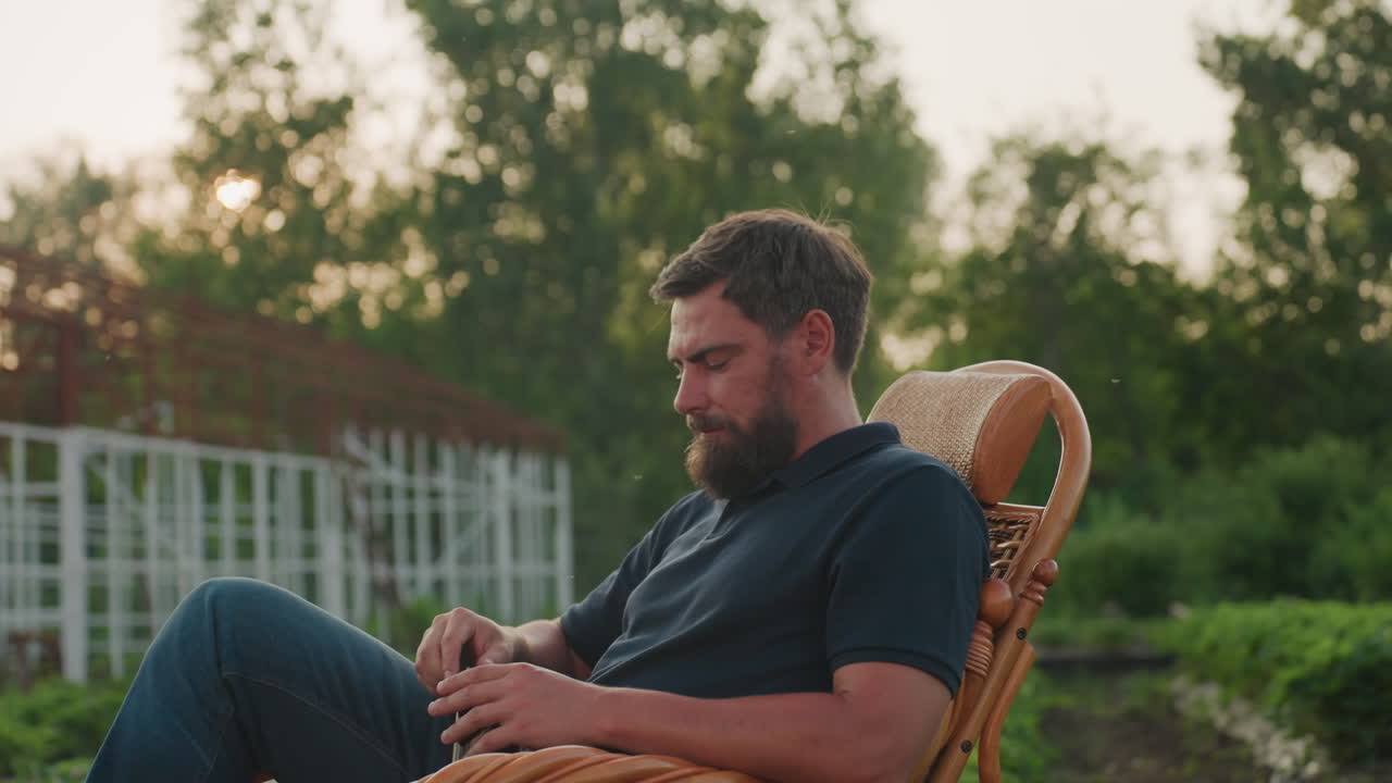 young man seated in wicker rocking chair in lush garden holds steaming herbal tea and savors quiet moment under golden sunset light, relaxed rural scene