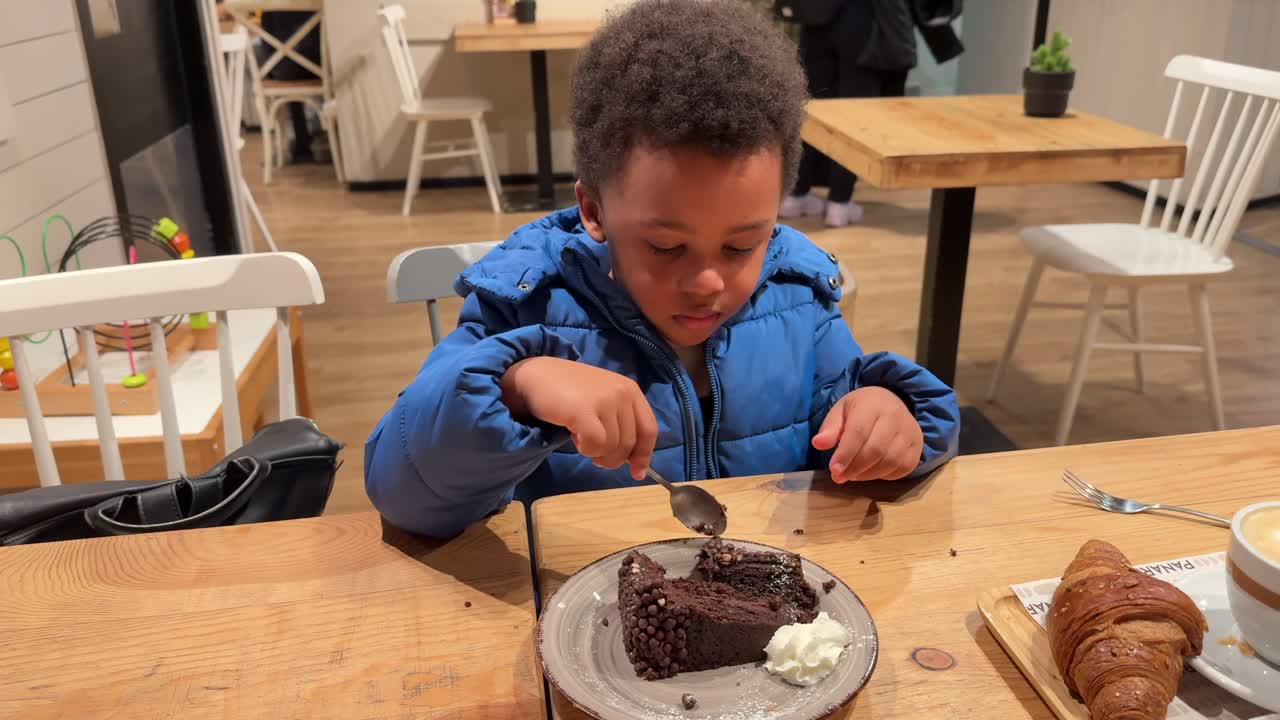 niño negro biracial glotón de 4 años comiendo un pastel de chocolate solo sentado en una mesa de madera dentro de una cafetería. lleva el cabello rizado y ropa de invierno. 4k