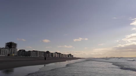 Runner-Running-On-The-Beach-In-North-Sea,-Knokke,-Belgium-With-Waves-Splashing-To-The-Sand-On-A-Sunrise---wide-drone-shot