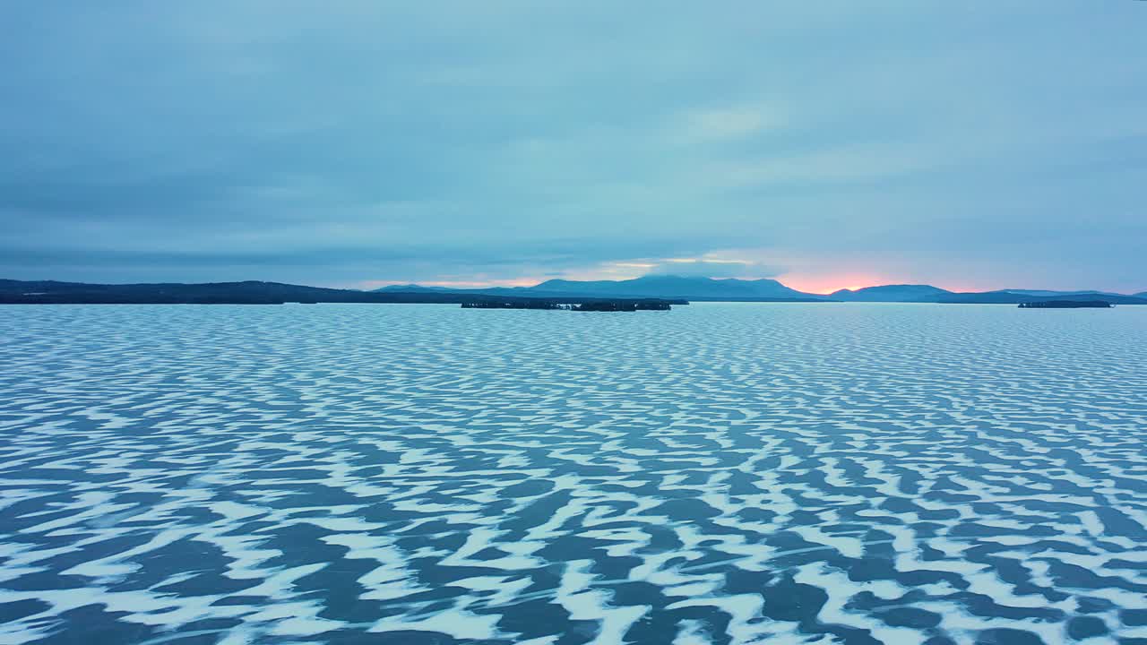 deslizamiento aéreo sobre un lago congelado con patrones nevados en el hielo y el color del amanecer asomando a través de la cordillera en el horizonte