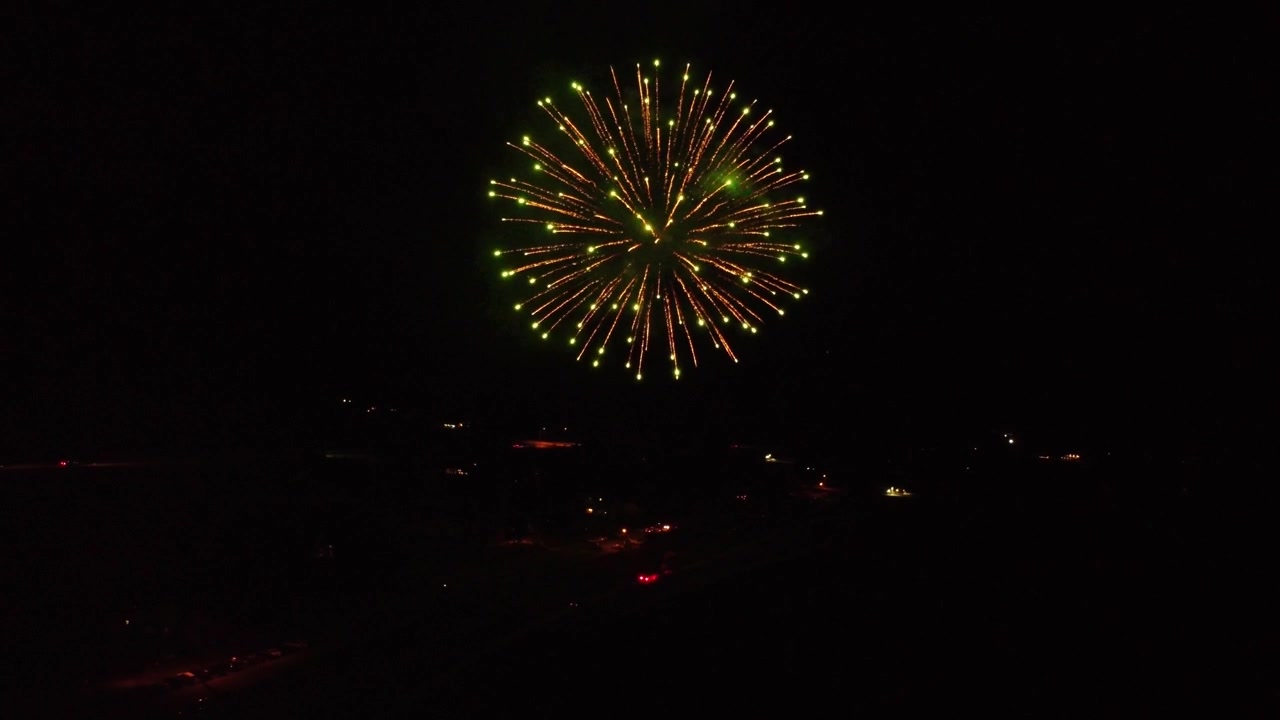 Fireworks exploding going off in the dark of night as drone is flying close to the show, then flys back to reveal lake with colorful explosive reflections in water