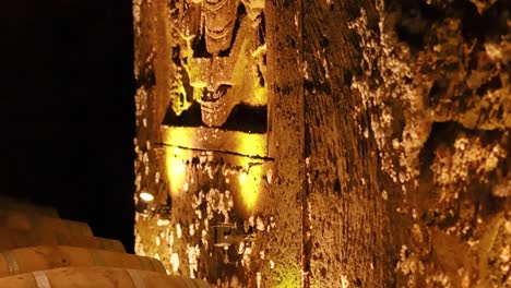 close-up of a textured stone wall with warm lighting and partial view of wine barrels.