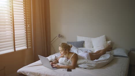 joven sensual con camiseta blanca y bragas acostada en la cama cerca de la computadora portátil