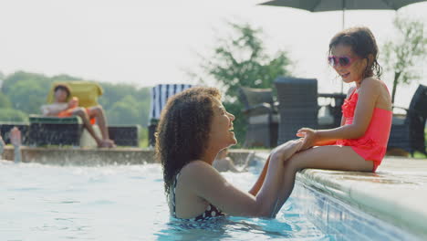 mother and daughter have fun in outdoor swimming pool on summer vacation with girl sitting on edge