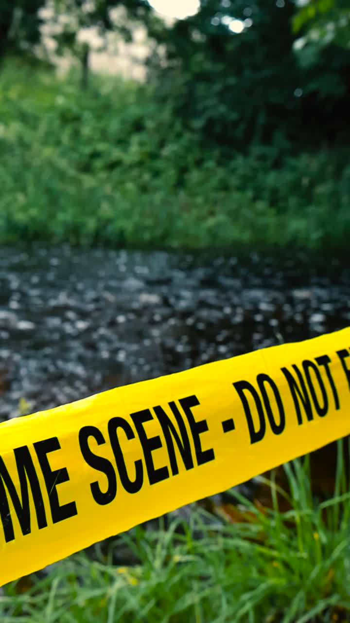 Vertically filmed footage of a dark fast flowing river with a yellow colored vibrant police crime scene tape do not enter ribbon pulled in front of the grassy shoreline at summer cloudy day