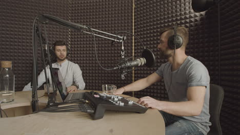 the bearded radio presenter turns down the volumen of the sound mixer while a young man next to him is talking in a radio recording studio