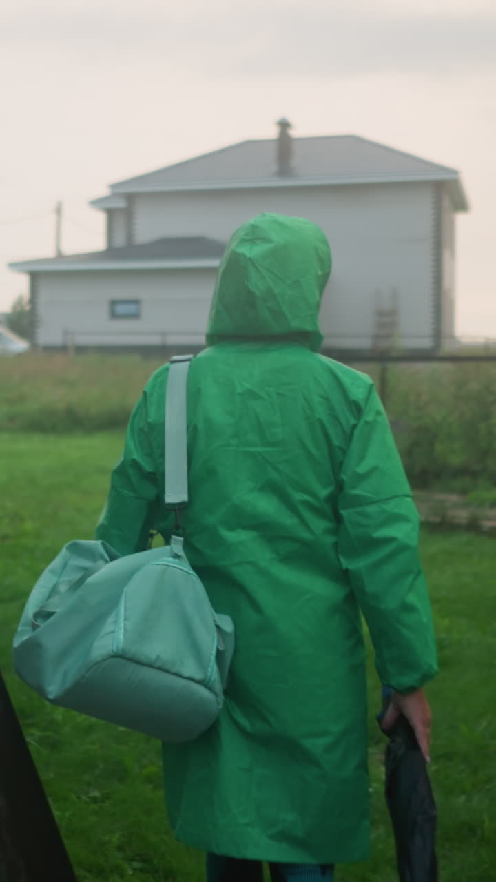 vista trasera de un individuo con una impermeable verde que lleva una bolsa de color menta y un paraguas mientras camina hacia un área de hierba, la escena incluye un banco oscilante, un edificio y un automóvil en la distancia