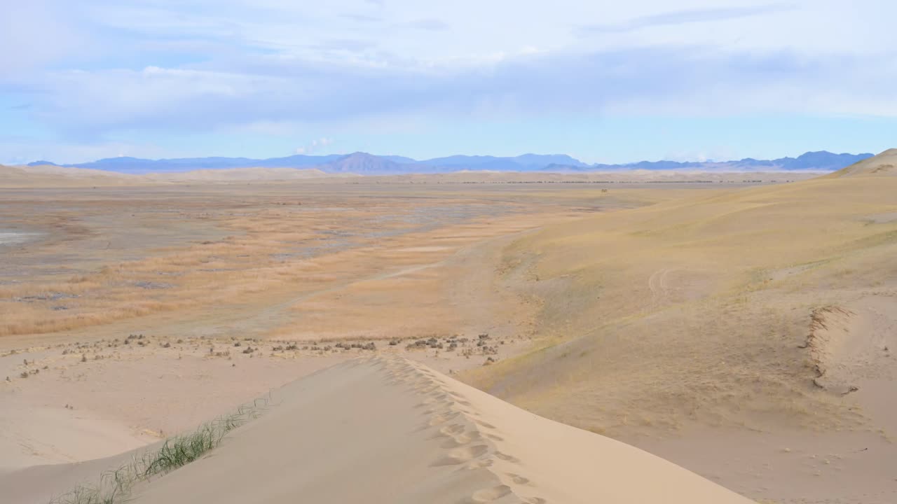 A trail of footprints leads along the crest of a sand dune, overlooking the vast, arid landscape of the Durgun Nuur desert. A scene of a lonely journey or exploration