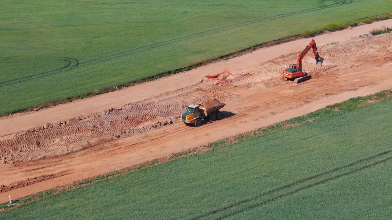 Ripresa dall'alto di macchinari pesanti che costruiscono la tangenziale occidentale di Stettino in Polonia. Un escavatore e un dumper lavorano su un sentiero sterrato attraverso campi verdi