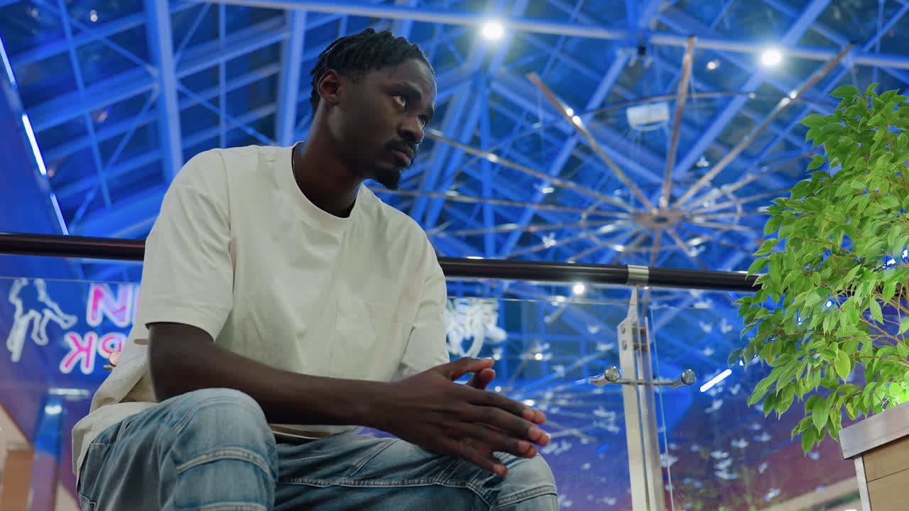 Music artist seated in modern shopping mall wearing white t-shirt and jeans tapping hand thoughtfully while waiting for someone, indoor scene with glass ceiling and greenery