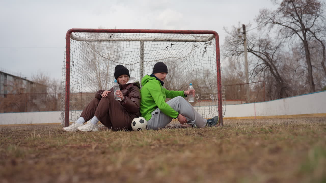 adolescentes blancos apoyados espalda con espalda en la portería, con una postura relajada, con un balón de fútbol a los pies, cielo nuboso y campo vacío, sonrisas discretas y chaquetas cálidas, encuentro nostálgico de fin de semana, vínculo juvenil e informal