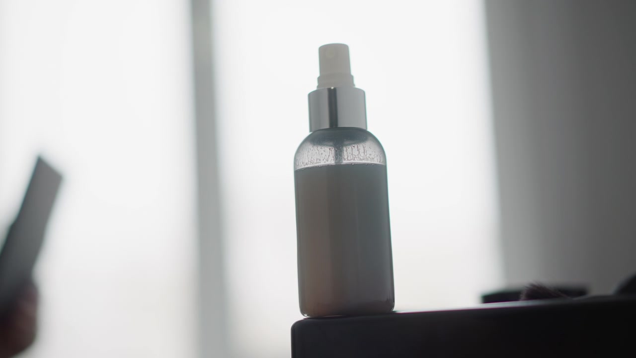 Close up of clear plastic spray bottle with white liquid and silver cap on black surface. Background features soft light blurred window view faint reflection calm atmosphere
