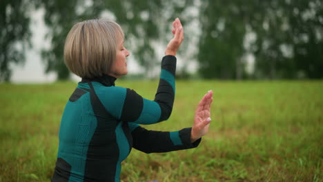 side view of middle-aged woman in green and black suit practicing yoga in cow face pose, seated outdoors in a peaceful grassy field under a cloudy sky, focusing on mindfulness and inner calm