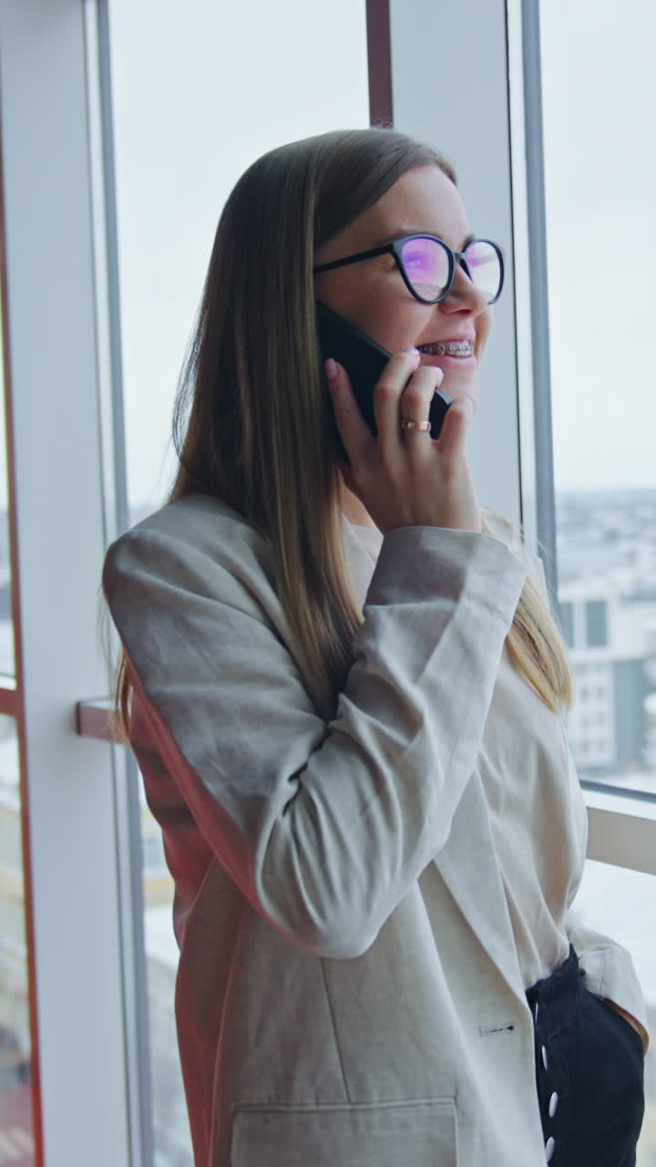 Attractive lady in glasses stands at the window speaking on the phone. Pleasant phone conversation in spacious office. Vertical video