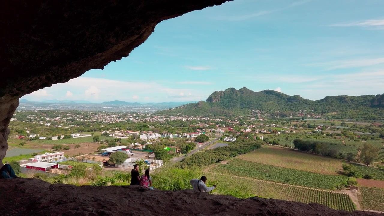 Vista da una grotta che si affaccia su una valle rurale con terreni agricoli, strada tortuosa e paesaggio montano aspro, circondata da scenari naturali di giorno a Morelos, Messico