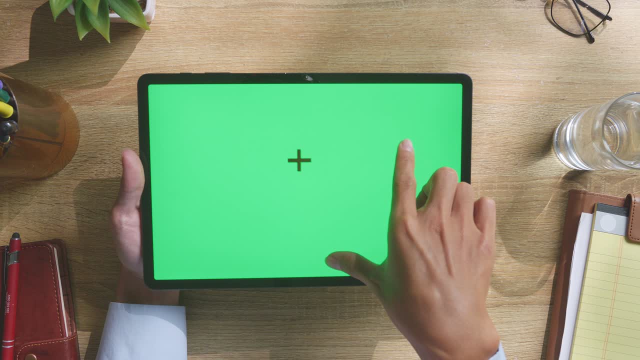 верхний вид ноутбука с макетным зеленым экраном chromakey на деревянном офисном столе рядом с ноутбуком с ручками, очками и стаканом воды.