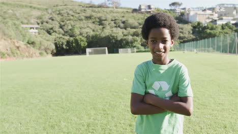 biracial boy stands confidently on a soccer field, wearing a green recycling t-shirt, with copy spac