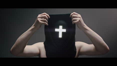 masked man with cross of light glowing on his face puts on a hood