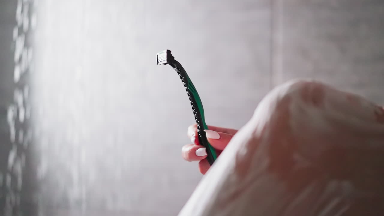 여성은 욕실 클로즈업에서 다리를 면도하기 위해 면도기를 들고 있습니다. 여자는 샤워 유에서 몸의 털을 제거하기 위해 현대적인 면도기를 보여줍니다. 피부 관리 및 아름다움