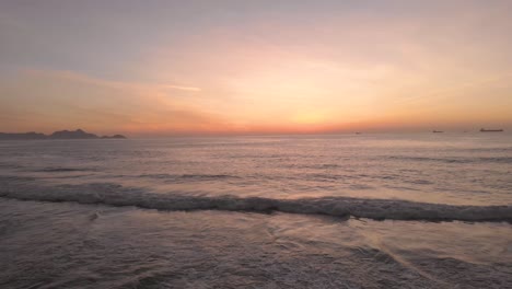 Aerial-backwards-movement-staying-away-from-an-incoming-wave-revealing-the-pristine-Devils-beach-in-Rio-de-Janeiro-just-before-sunrise-with-the-sky-glowing-with-bright-yellow,-orange-and-magenta